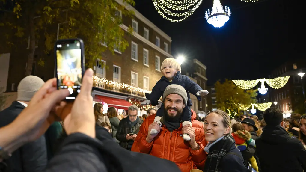 A man with a children on his shoulders has his photo taken by woman holding up a mobile phone